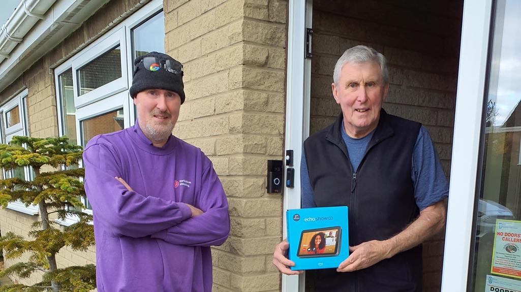 Man standing at his front door holding smart home technology hub box, with another man dressed in Yorkshire Housing work clothes
