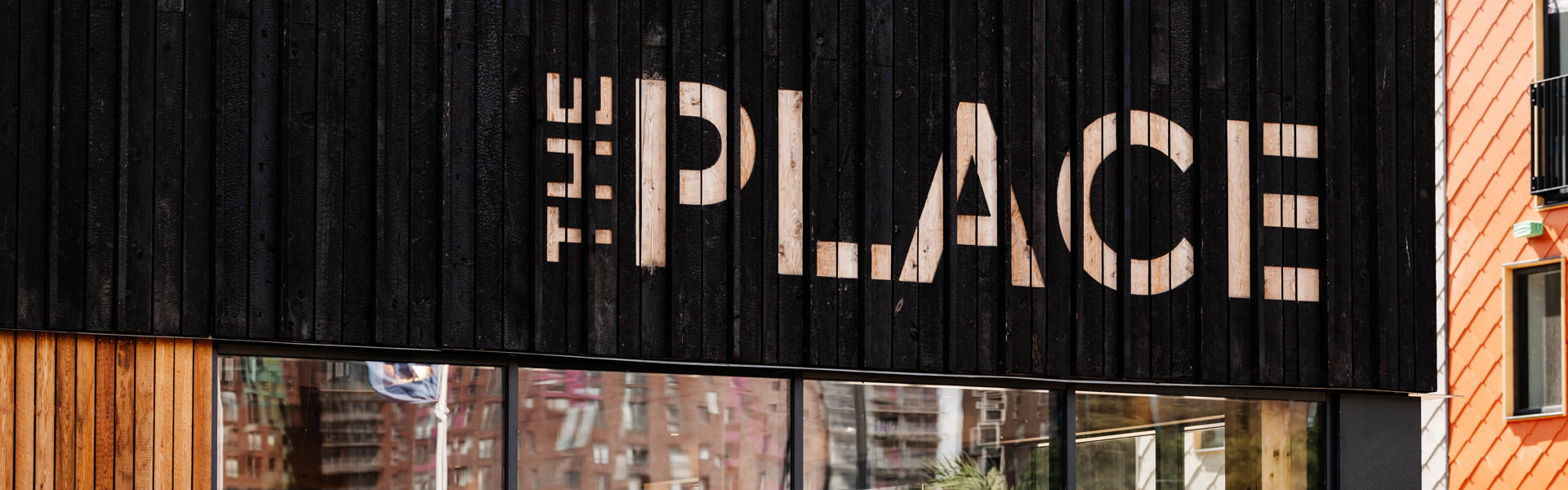 Photograph of the outside of The Place, showing a large glass wall with black wood above it with The Place spelled out in pale wood. 