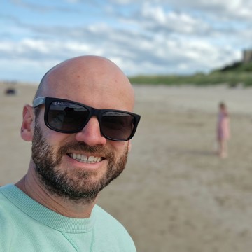 Rob wearing a mint green t-shirt smiling at the camera on the beach with his sunnies on 