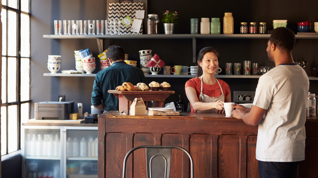 A photograph of a coffee shop with someone behind the counter smiling while serving a customer. 