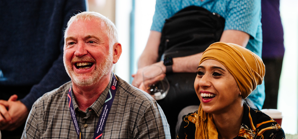 Man and woman sat next to each other laughing. The man is wearing a checked shirt and the woman is wearing a rope print shirt and mustard yellow headscarf. 