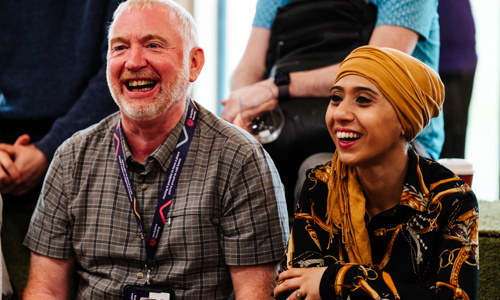 Man and woman sat next to each other laughing. The man is wearing a checked shirt and the woman is wearing a rope print shirt and mustard yellow headscarf.