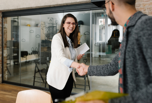 Woman in a white blazer and light blue shirt shaking the hand of a man wearing a more casual grey top. Both are smiling in a modern office.