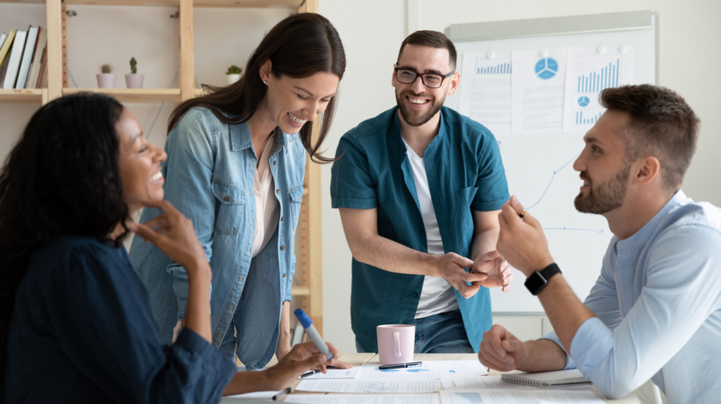 Four people in a work environment stood and sat round a table smiling an laughing