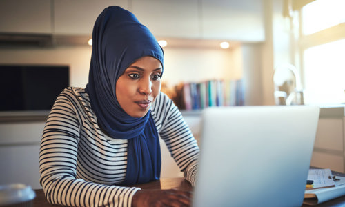 Women working at home, typing on a laptop while wearing a navy hijab and navy and white striped top.