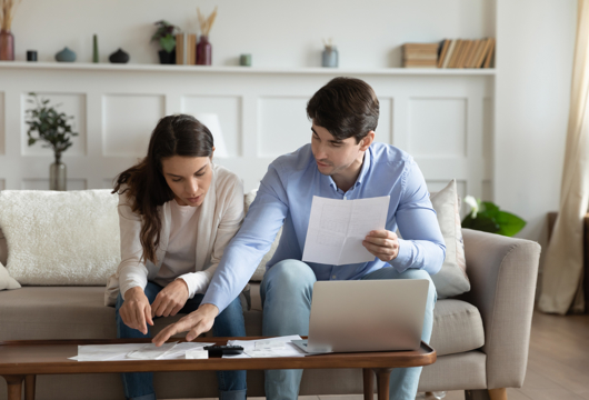 Photo of a couple sat on a sofa looking at papers together on the coffee table infront of them