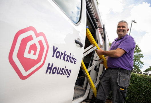 Passenger side of a white van with a Yorkshire Housing logo decal on the open sliding van door. A man is taking a ladder out of the van and looking and smiling at the camera.