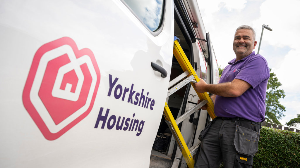 Passenger side of a white van with a Yorkshire Housing logo decal on the open sliding van door. A man is taking a ladder out of the van and looking and smiling at the camera.