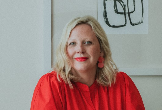 A photo of Catherine Asta sat at a table with her hands crossed and smiling at the camera.