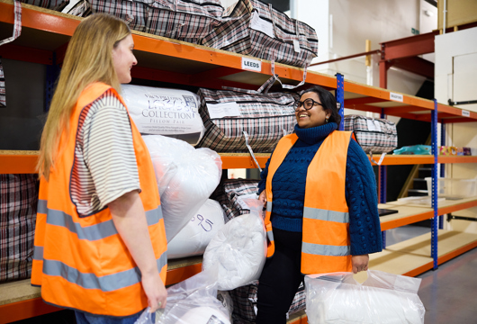 Two volunteers in the Zarach warehouse holding pillows in front of shelves of pillows
