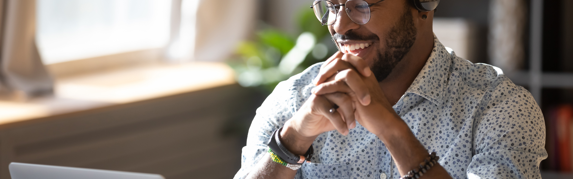 Man wearing a patterned shirt and glasses smiling at his computer, wearing a headset. 